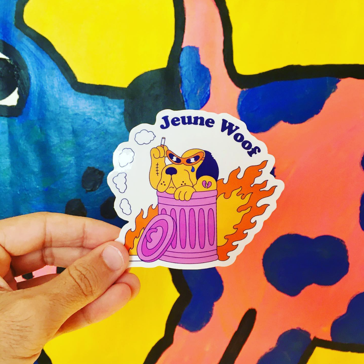 Hand holding a glossy vinyl sticker of a cartoon dog in a flaming trash can with "Jeune Woof" text, against a colorful abstract background.