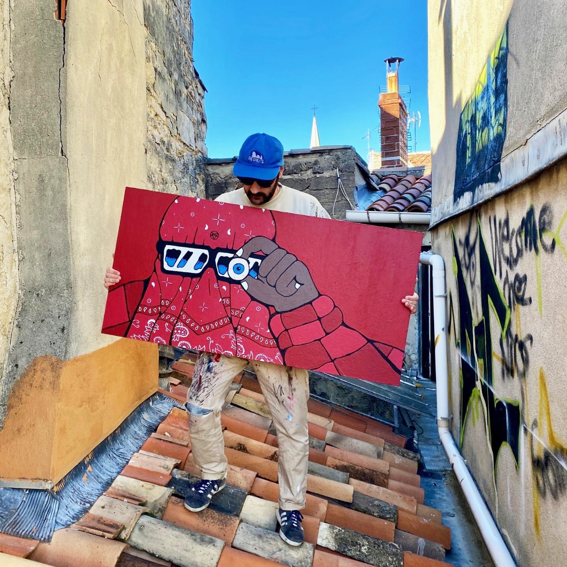 Artist Jeune Woof holding a red artwork of a figure with 3D glasses, standing on a rooftop with graffiti and a clear blue sky in the background.
