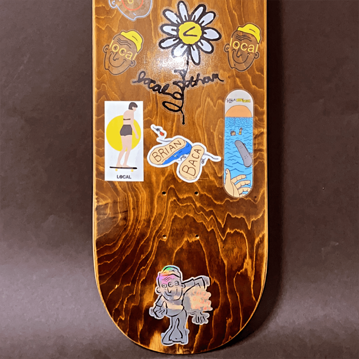 Skateboard deck with various colorful stickers, including cartoons, a flower, and text, on a wooden background.