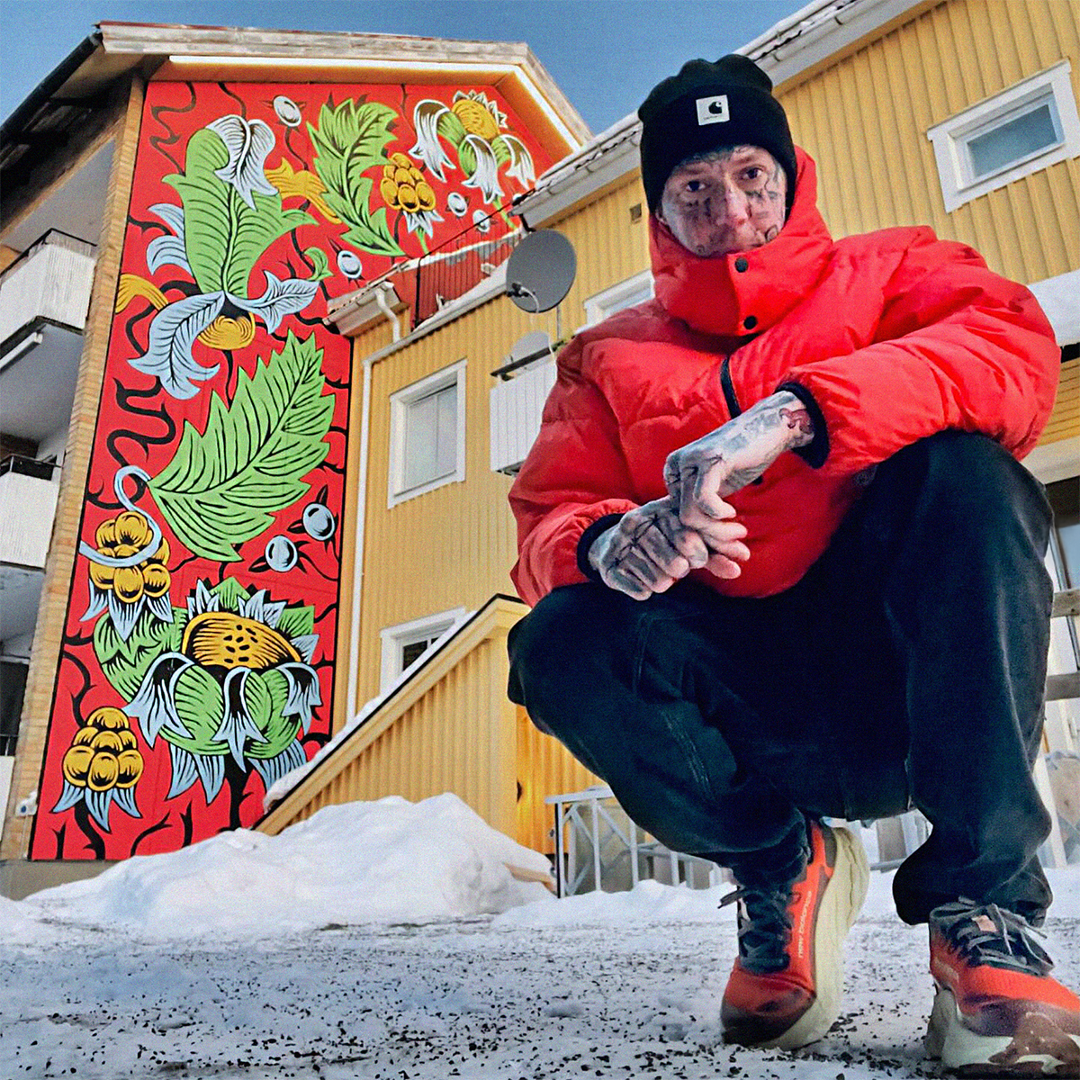 Marc UA in a red jacket and black beanie squats in front of a colorful mural on a snowy street.