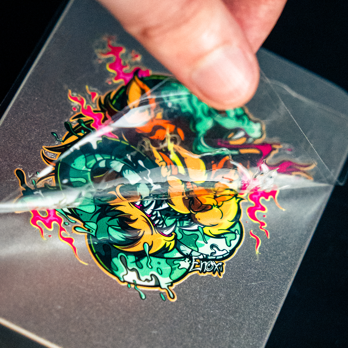 A hand peeling a vibrant, colorful transfer sticker with abstract graffiti-style art, featuring bold shapes and bright splashes of pink and green.
