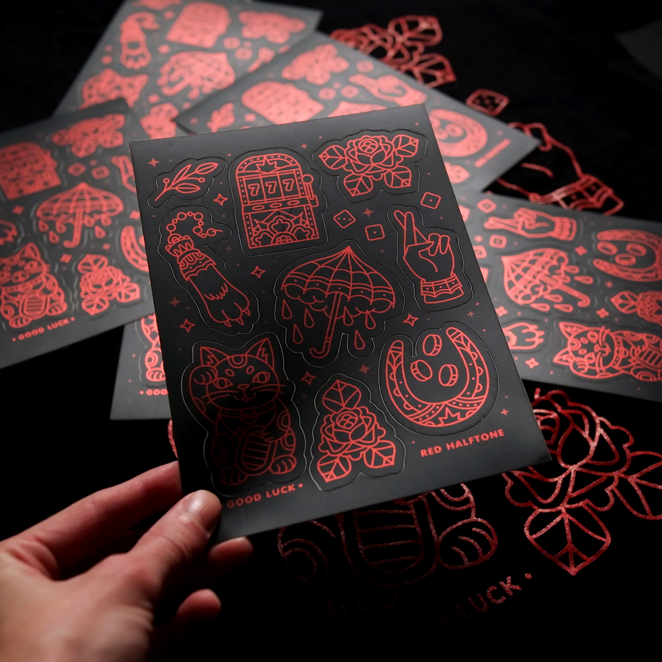 Hand holding a custom sticker sheet in matte mirror with red illustrations, including a lucky cat, dice, flowers, and other symbols.