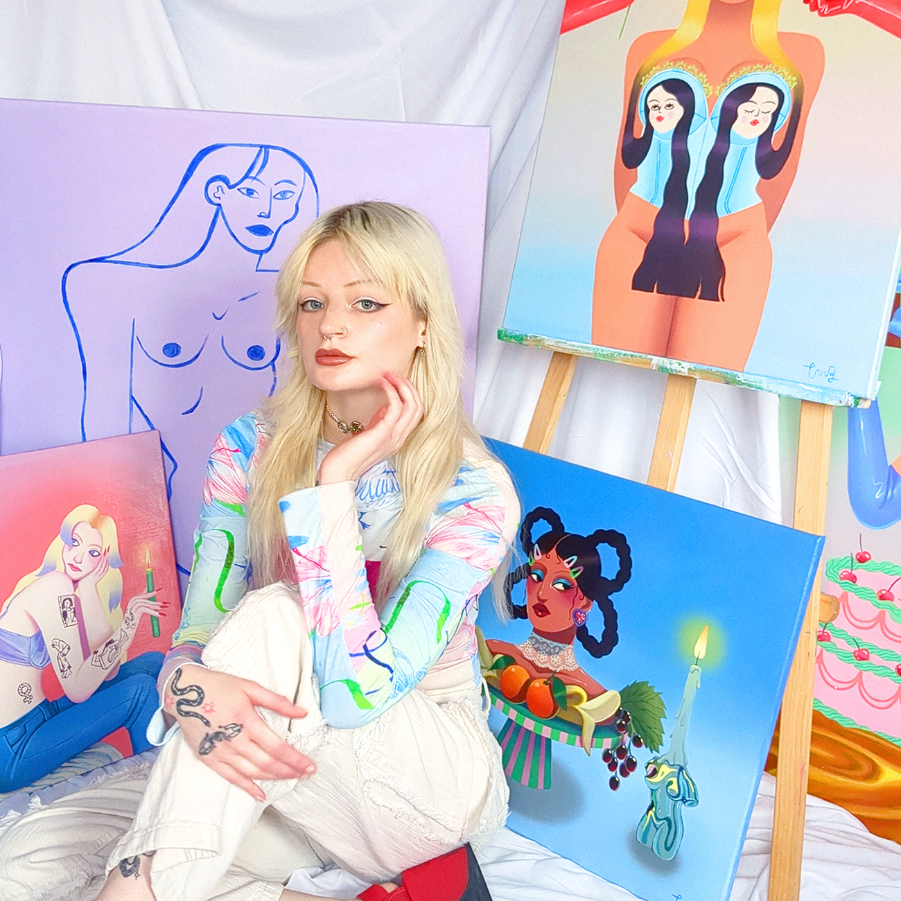 Artist sitting among colorful paintings, featuring abstract figures and vibrant designs, in a bright studio setting.