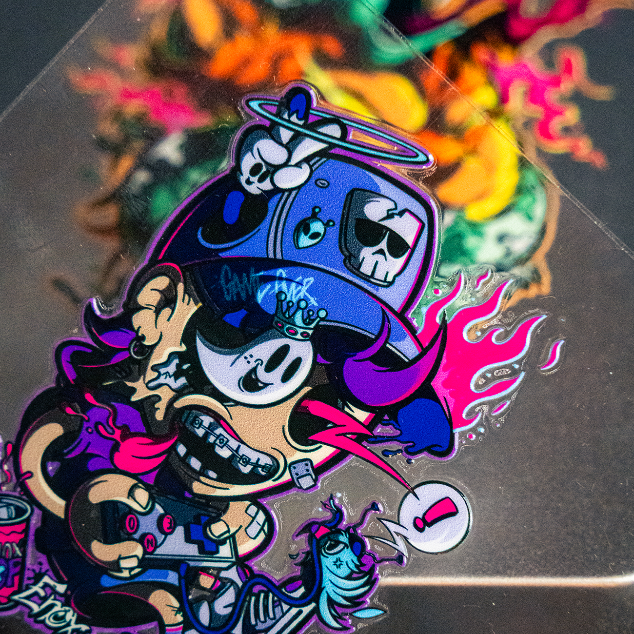 Colorful graffiti-style art of a cartoon transfer sticker with a cap, skull motifs, and vibrant colors, set against a blurred background.