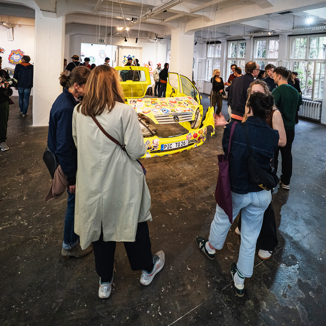 People looking at a car at an exhibition hanging from a ceiling, covered in different shaped and colored bumper stickers.