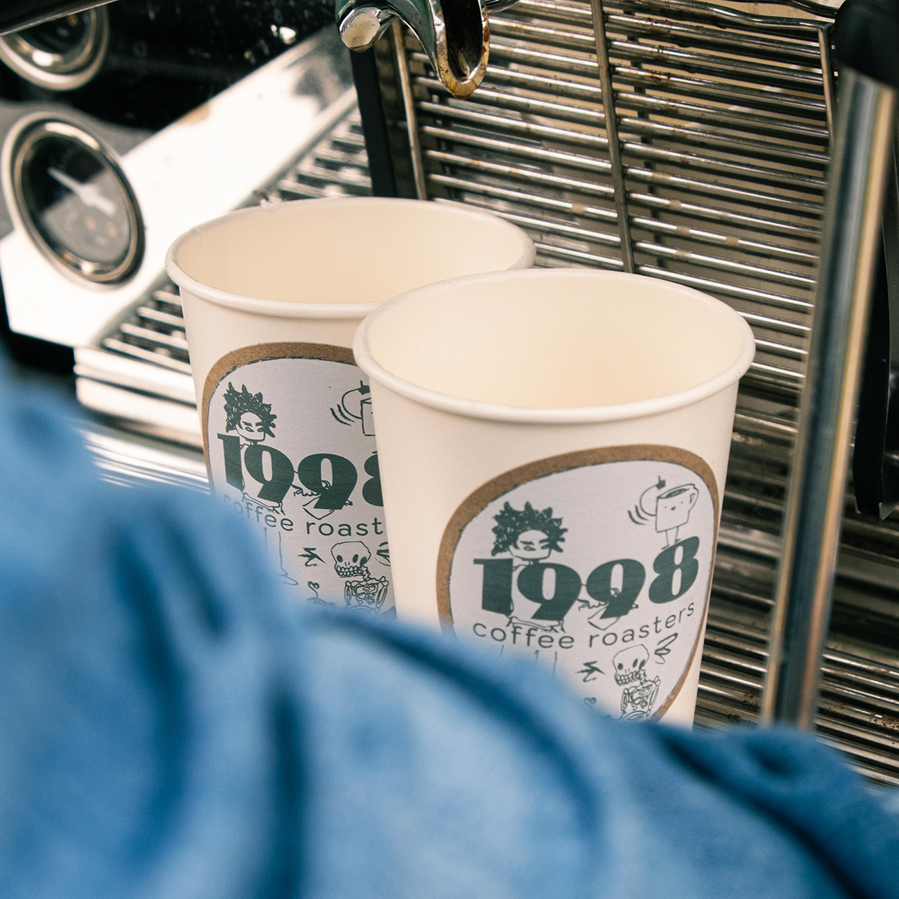 Two paper coffee cups with "1998 coffee roasters" kraft paper stickers placed near a metallic coffee machine, partially obscured by a blue fabric.