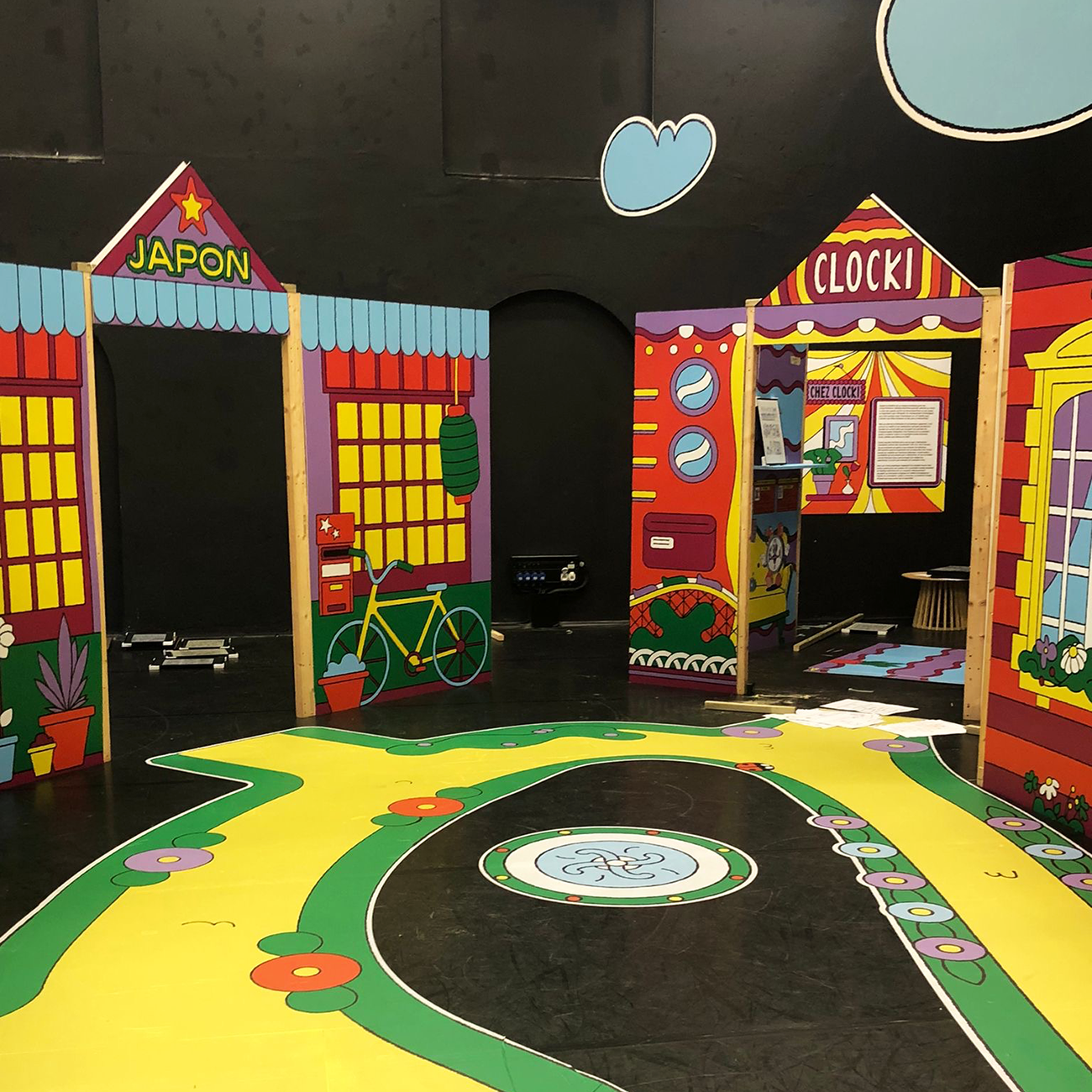 Colorful indoor play area with cartoon-style building stickers labeled "Japon" and "Clock," featuring paths, a bicycle, and various playful decorations.