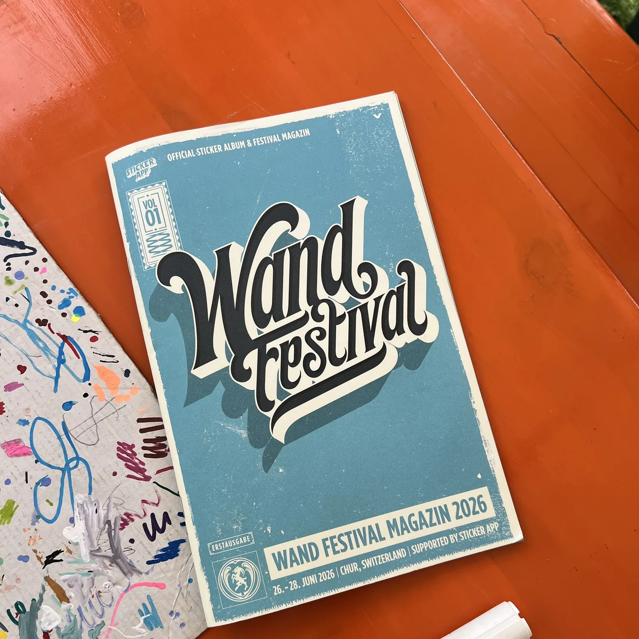 Officieel stickeralbum en festivalmagazine van Wand Festival 2026, gesteund door StickerApp, naast een verfstift op een oranje tafel