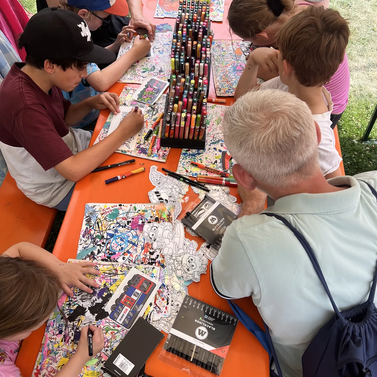 Kinderen kleuren contourgesneden stickers van Brainfart in rond een rek met verfstiften tijdens een festivalworkshop buiten