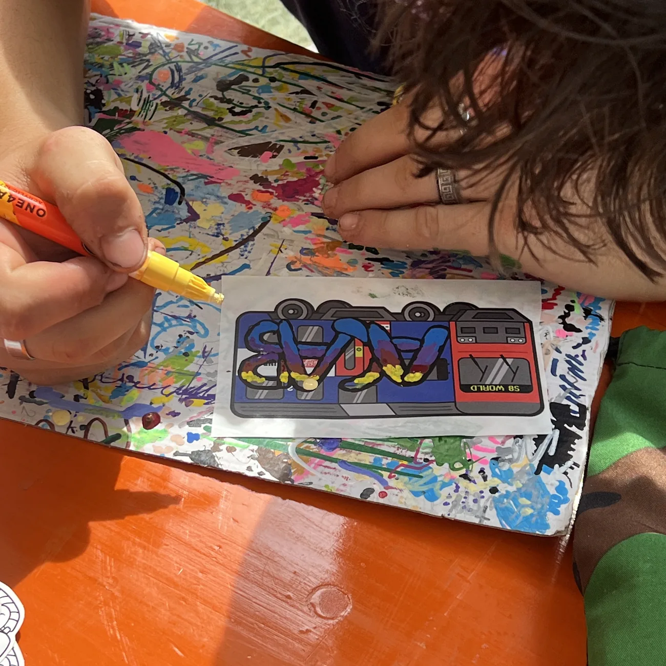 Een hand kleurt een etiket met de S8 World-trein in met een gele verfstift tijdens de workshop van Brainfart x StickerApp