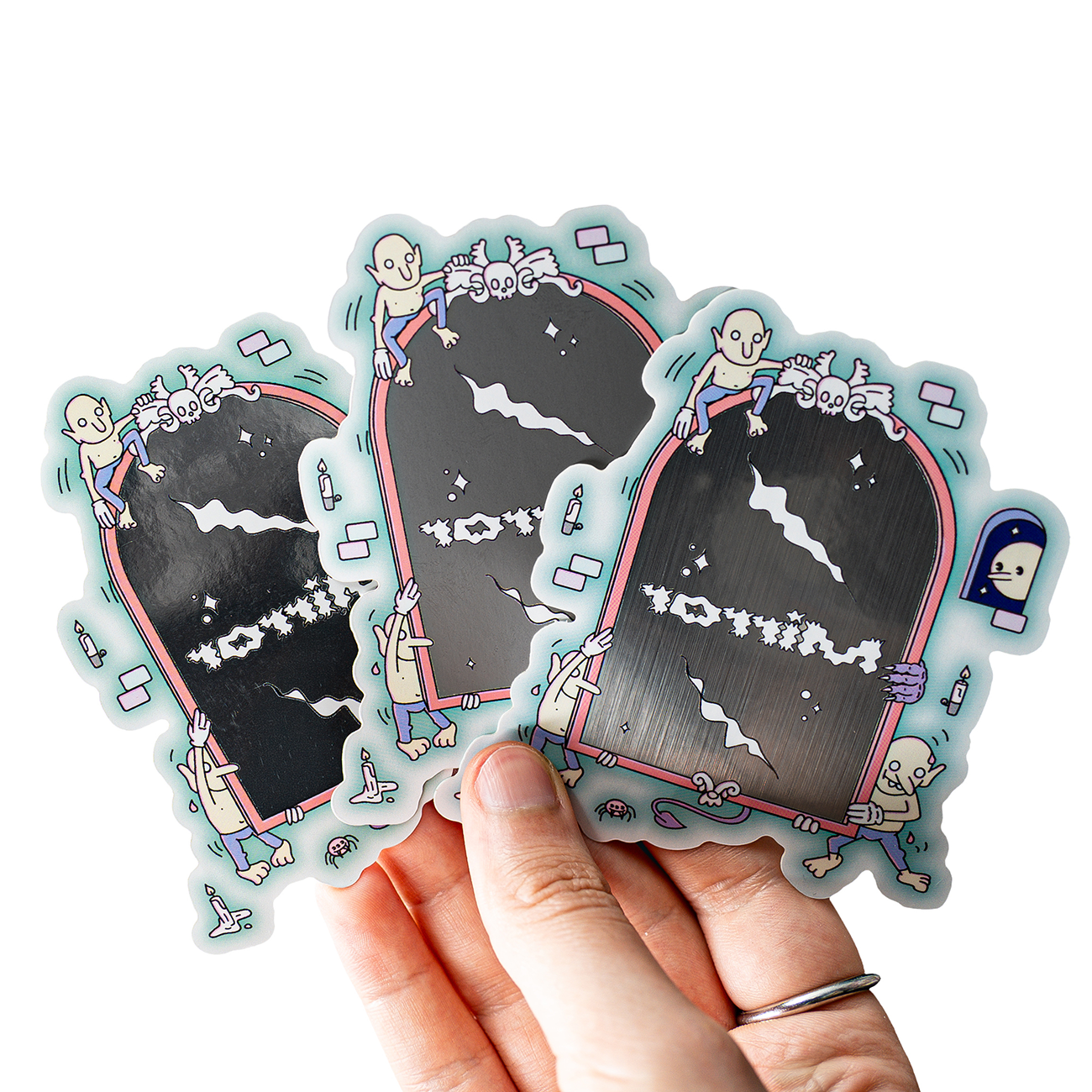 Hand holding three metallic stickers featuring cartoon skeletons climbing on oxidized steel mirrors with playful designs.