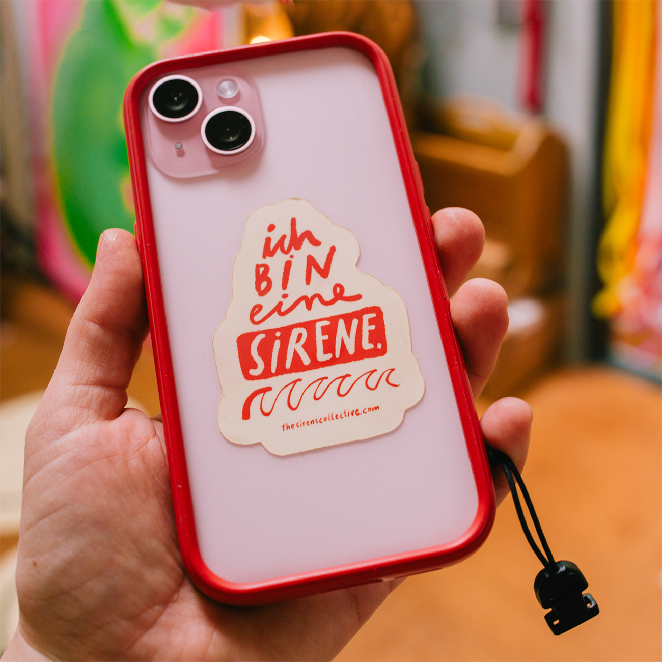 Hand holding a smartphone with a red case, featuring a vinyl die-cut sticker that reads "ich BIN eine SIRENE" in red text.