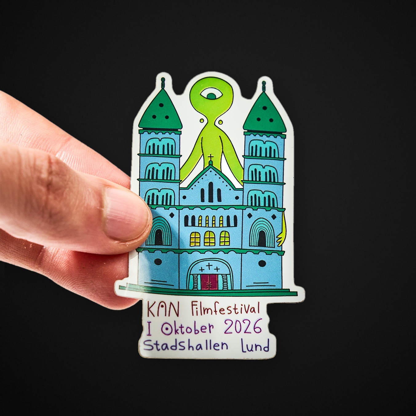 Hand met een contourgesneden sticker van een kathedraal met een groen eenogig wezentje, met de tekst “KAN Filmfestival I Oktober 2026 Stadshallen Lund”