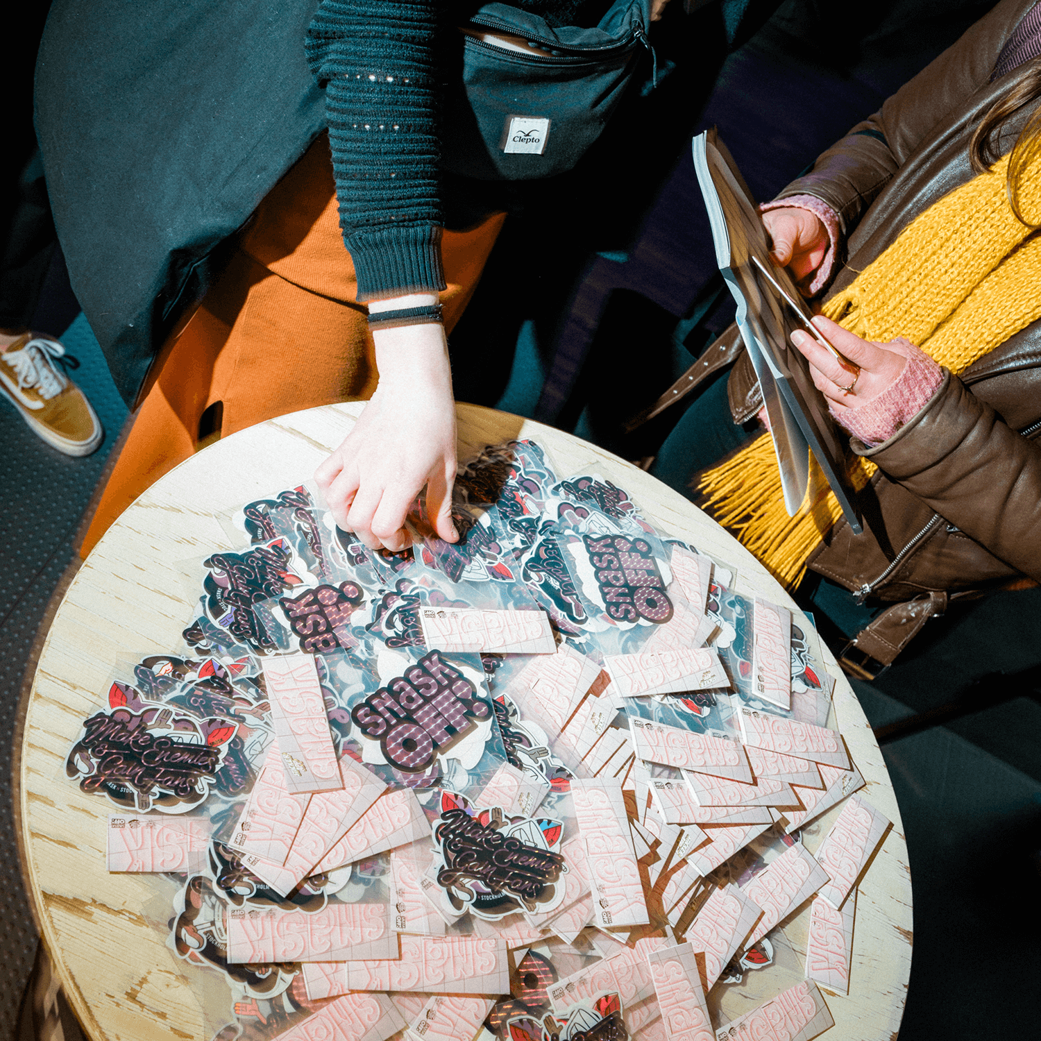 Two people stand by a round table covered with colorful sticker packs including different die cut stickers.