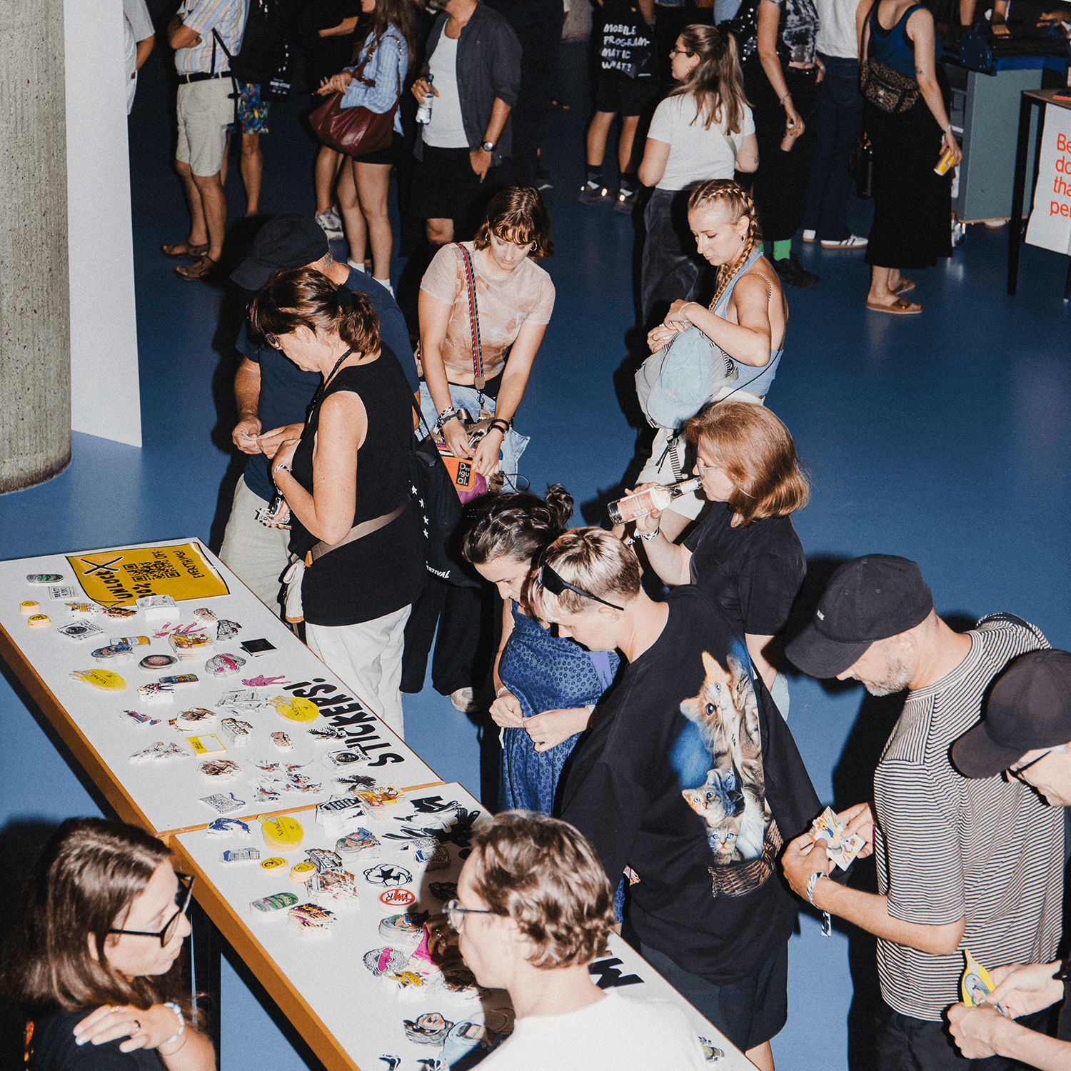 People gathered around a table with stickers, engaging in conversation and browsing stickers in a brightly lit room.