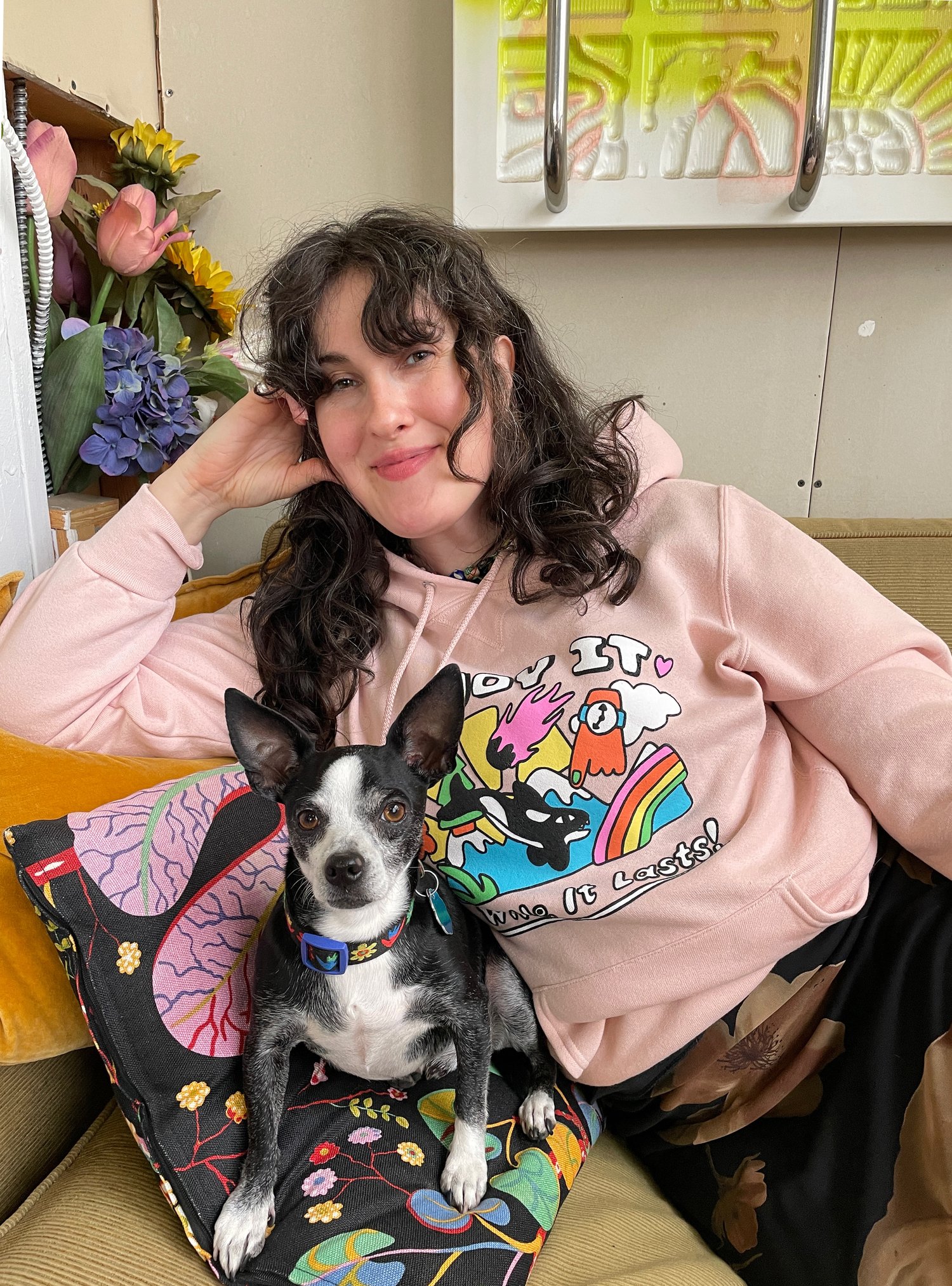Lachende Grace Miceli in een kleurrijke hoodie naast haar hond die op een kussen ligt