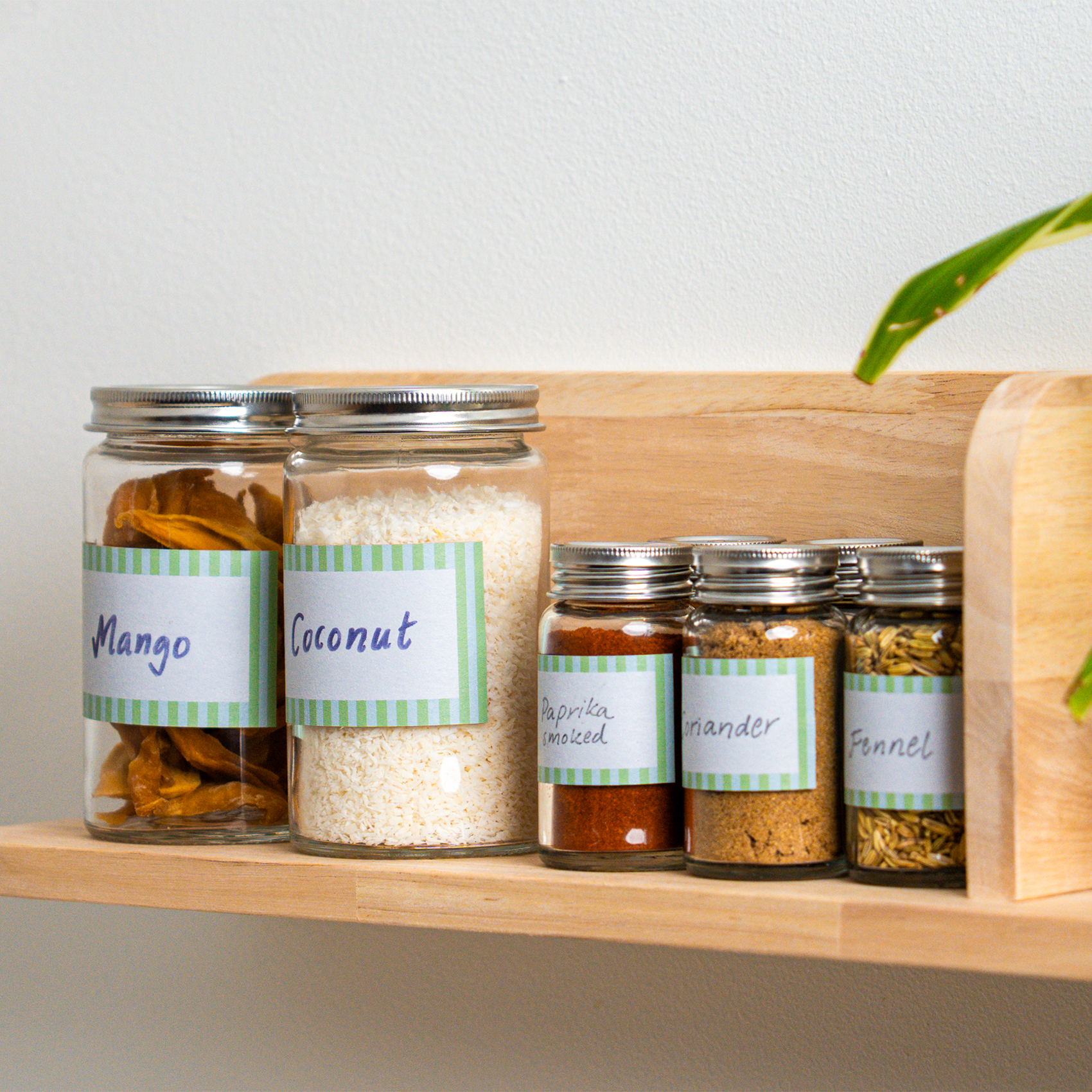 Glazen potten met handgeschreven etiketten op een houten keukenplank. In de potten zitten gedroogde mango, kokos en verschillende kruiden, waaronder gerookte paprika, koriander en venkelzaad.