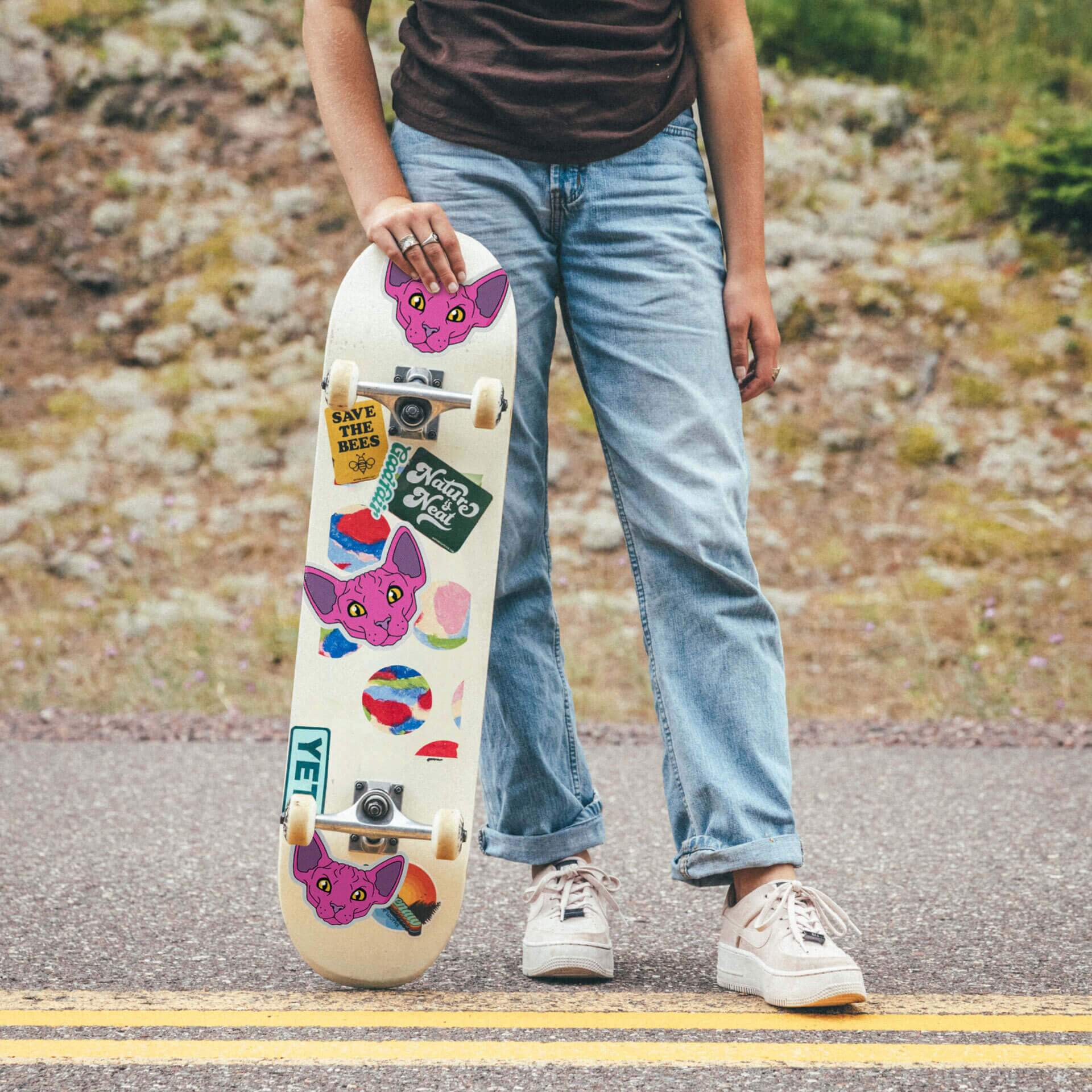 Person standing on a road holding a skateboard with colorful stickers, including a pink cat and various designs.