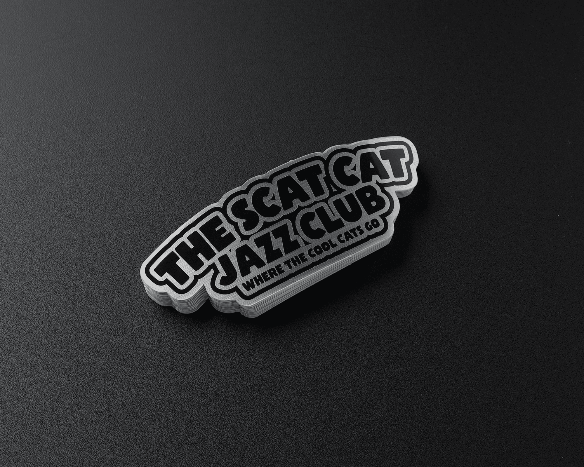 Stack of brushed aluminum stickers on a dark surface with bold text: "The Scat Cat Jazz Club - Where the Cool Cats Go."