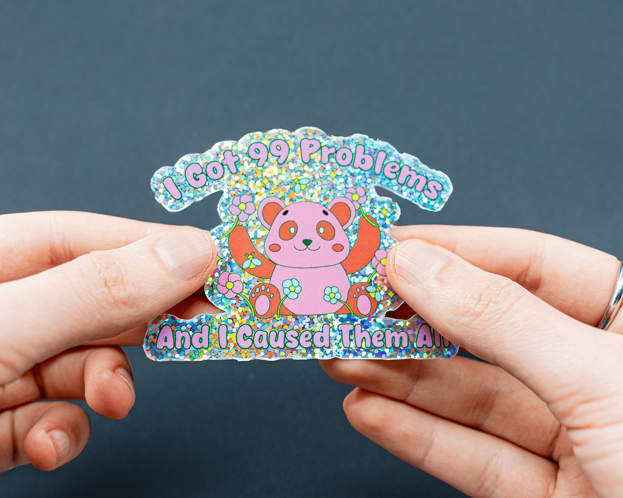 Hands holding a glitter sticker with a cute panda and the text "I Got 99 Problems And I Caused Them All" on a dark background.