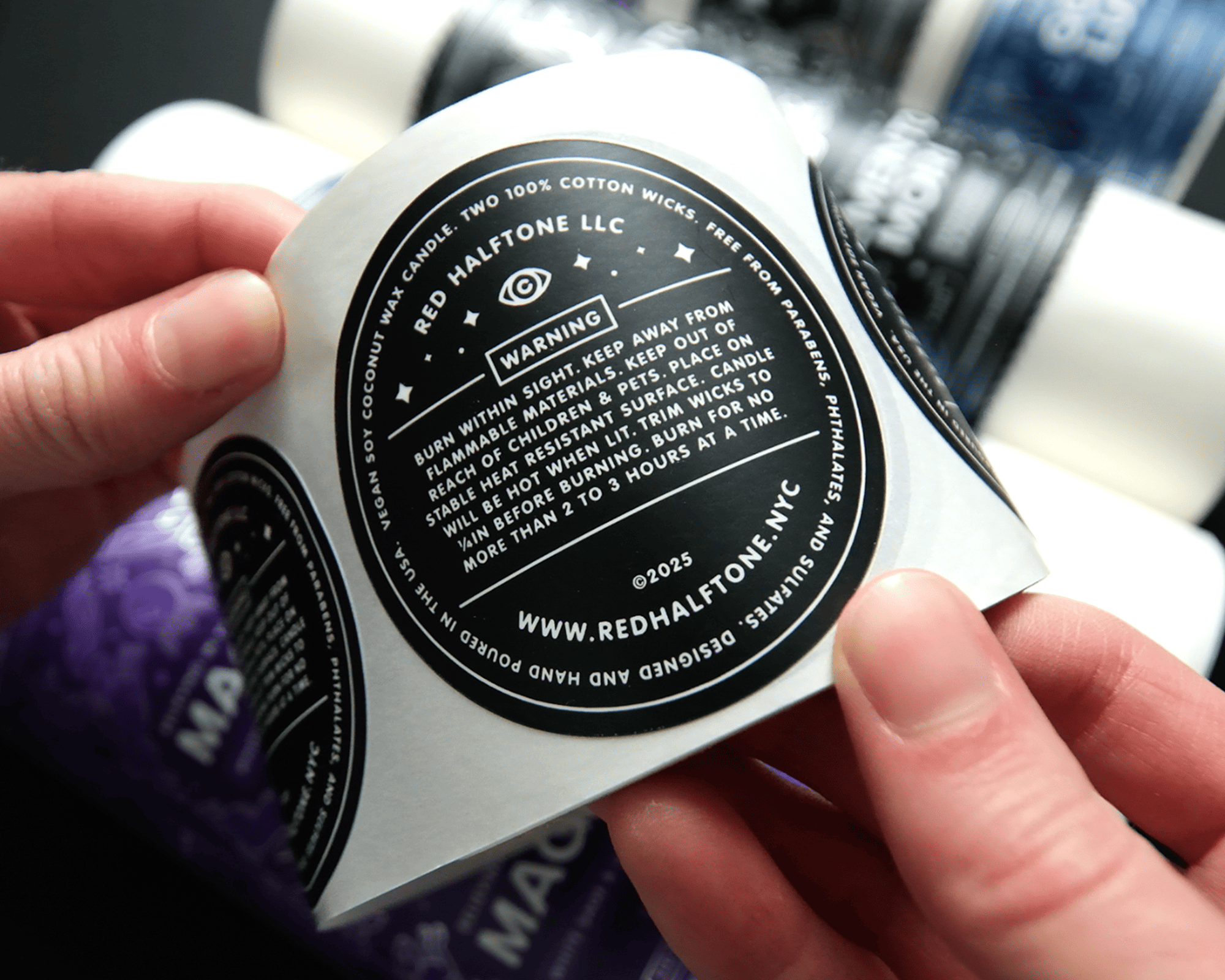 Hands holding a round candle label with safety instructions and a website URL, against a blurred background of more labels.