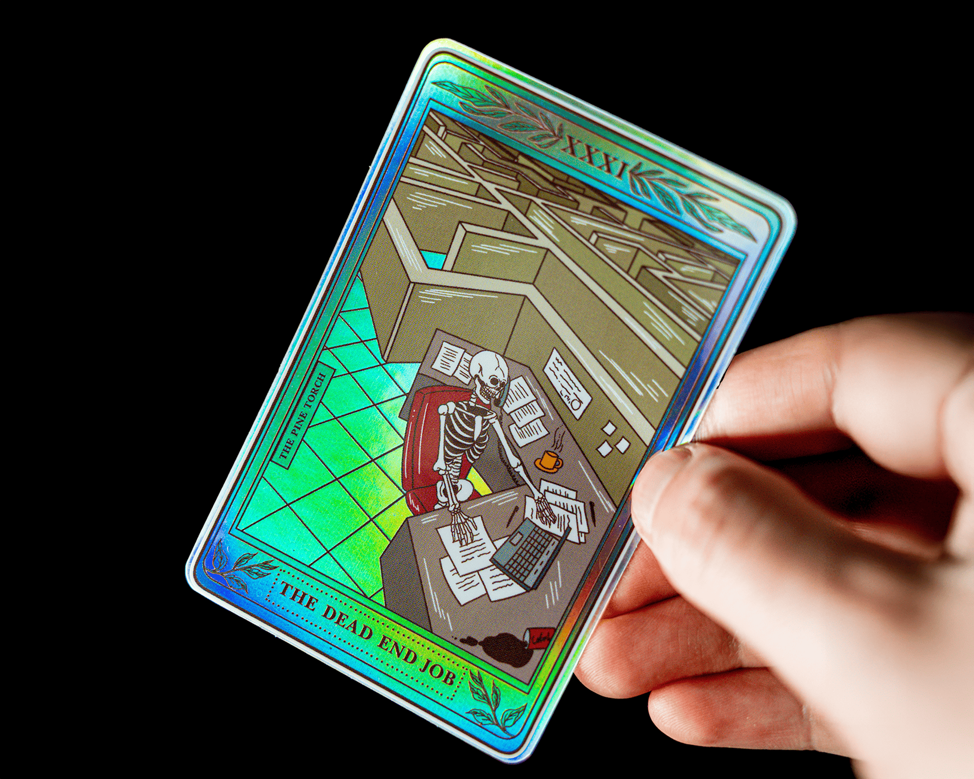 A hand holds a holographic sticker depicting a skeleton at a desk in a cubicle, titled "The Dead End Job."