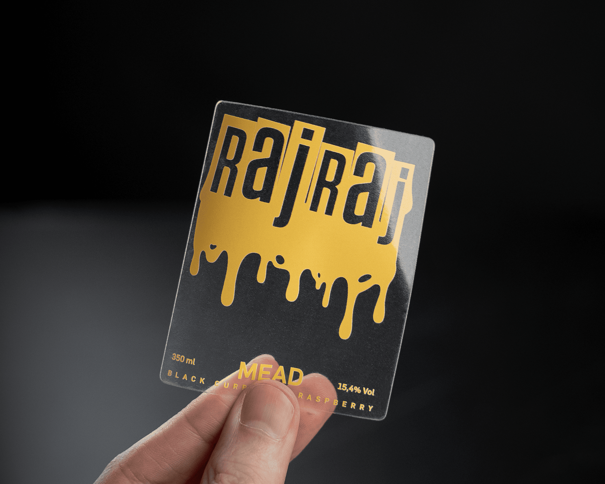 Hand holding a transparent label with "Raj Raj" in bold letters and dripping gold effect, featuring details about black currant raspberry mead.