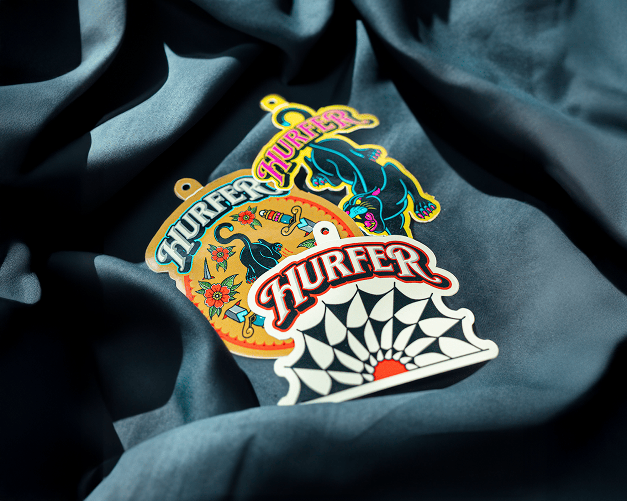 Three colorful hang tags with "HURFER" text, featuring a cat, a floral design, and a geometric pattern, placed on a dark fabric.