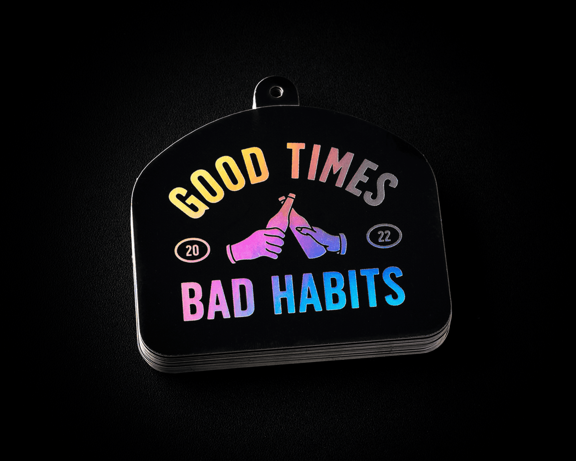 A hang tag with "Good Times Bad Habits" in colorful text, featuring two hands clinking bottles, and the numbers "20" and "22."
