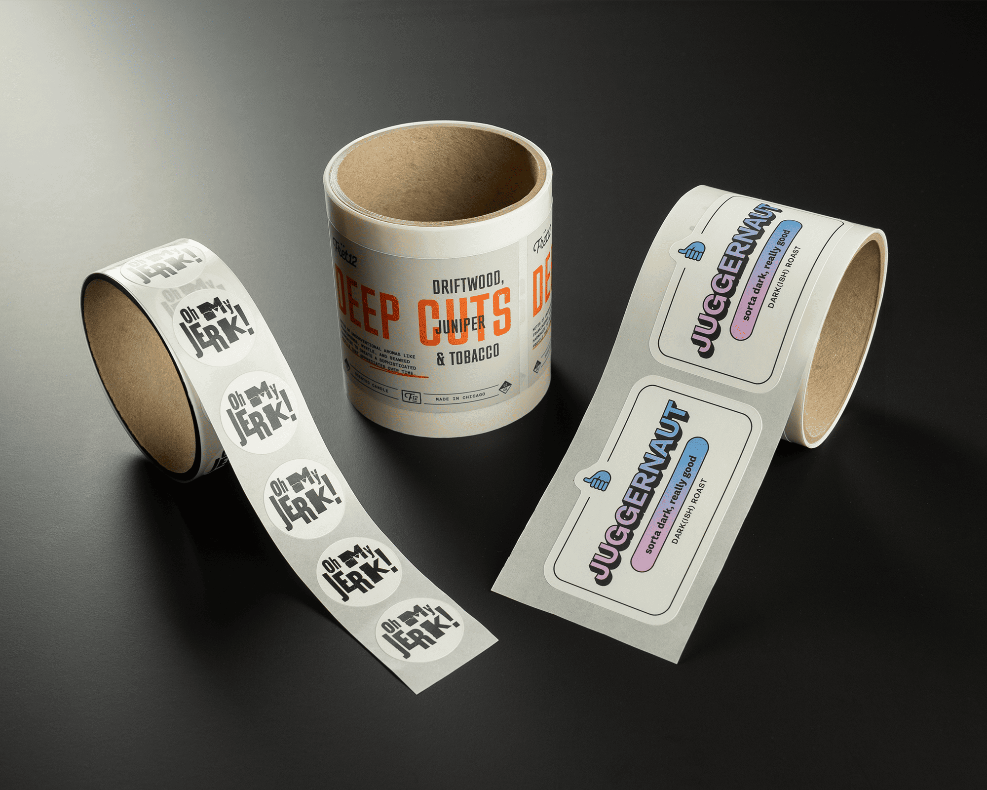 Three rolls of custom labels on a dark surface, featuring designs with text: "Oh My Jerk!" and "Juggernaut."