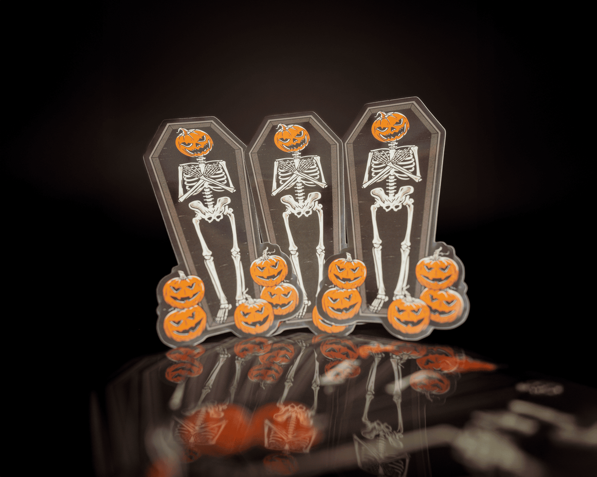 Three skeleton stickers with jack-o'-lantern heads and pumpkins, set against a dark background with reflections.