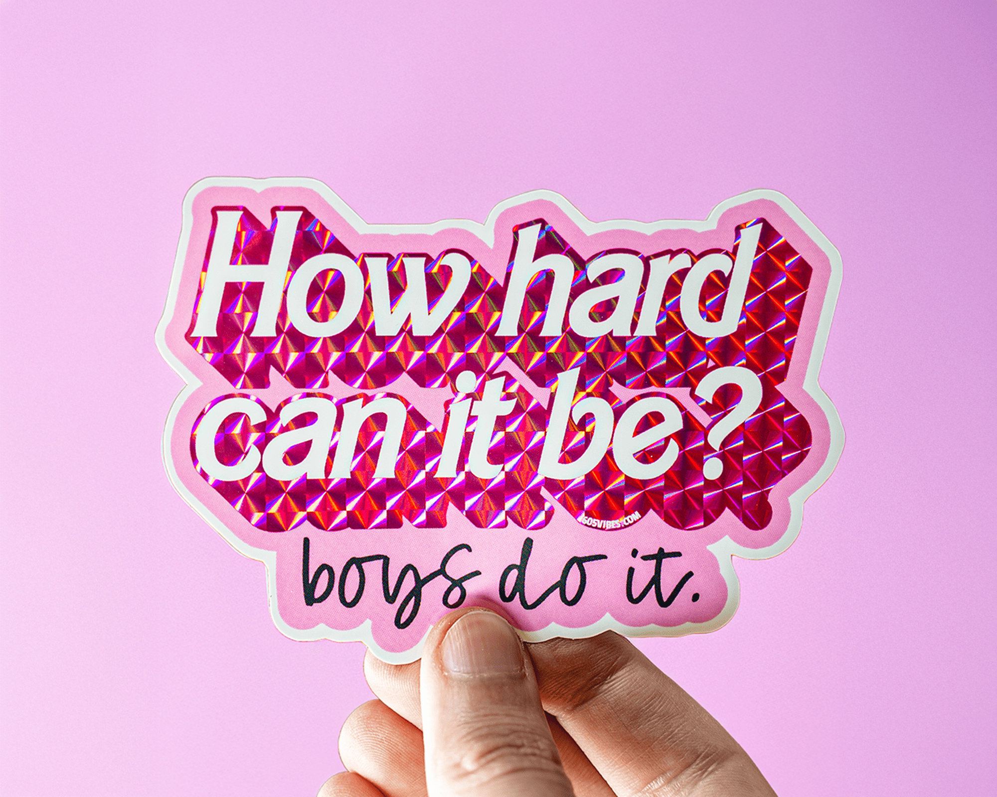 Hand holding a prismatic sticker with the text "How hard can it be? boys do it." on a pink background.