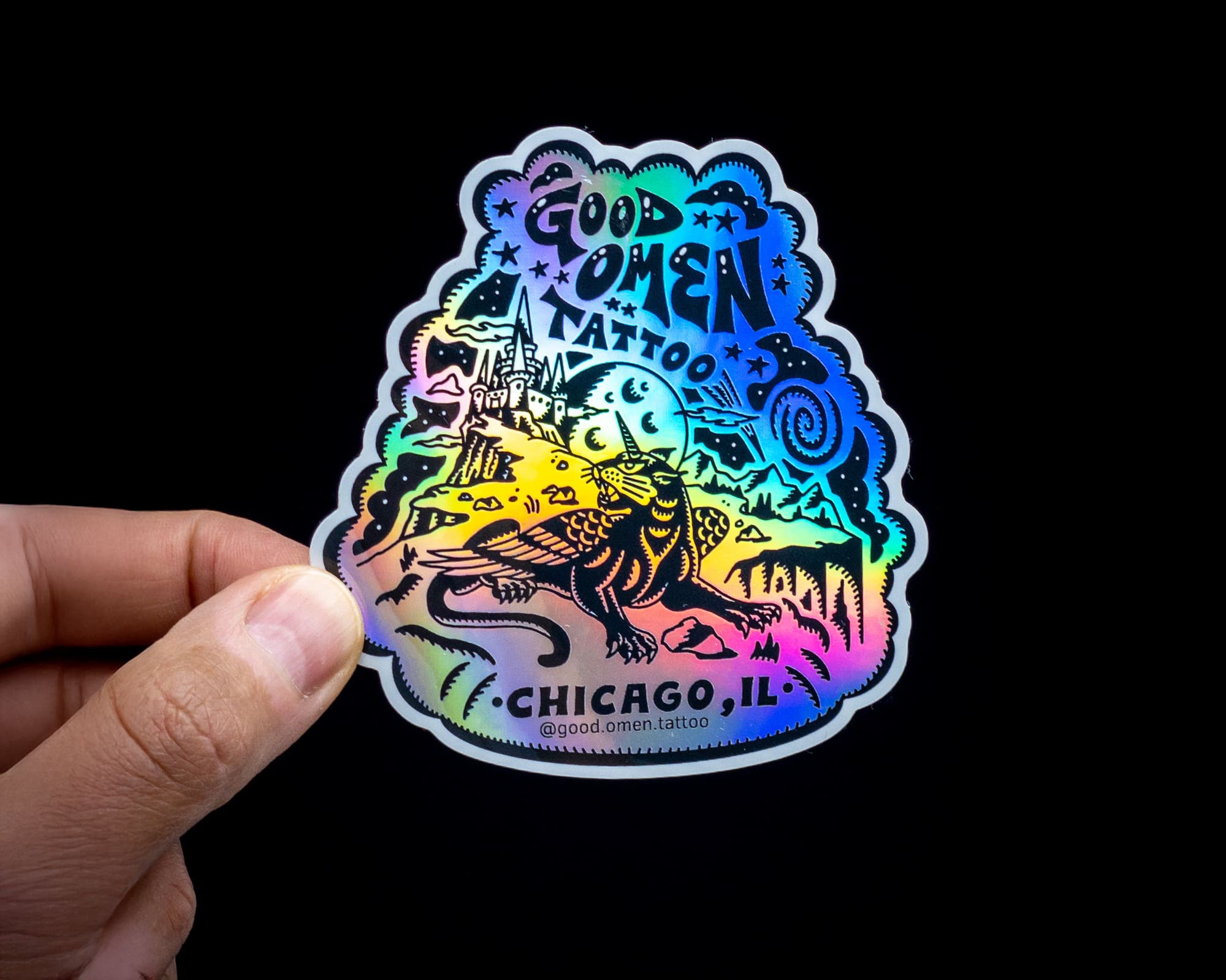 A hand holds a holographic sticker with a dragon, castle, and "Good Omen Tattoo, Chicago, IL" text against a black background.