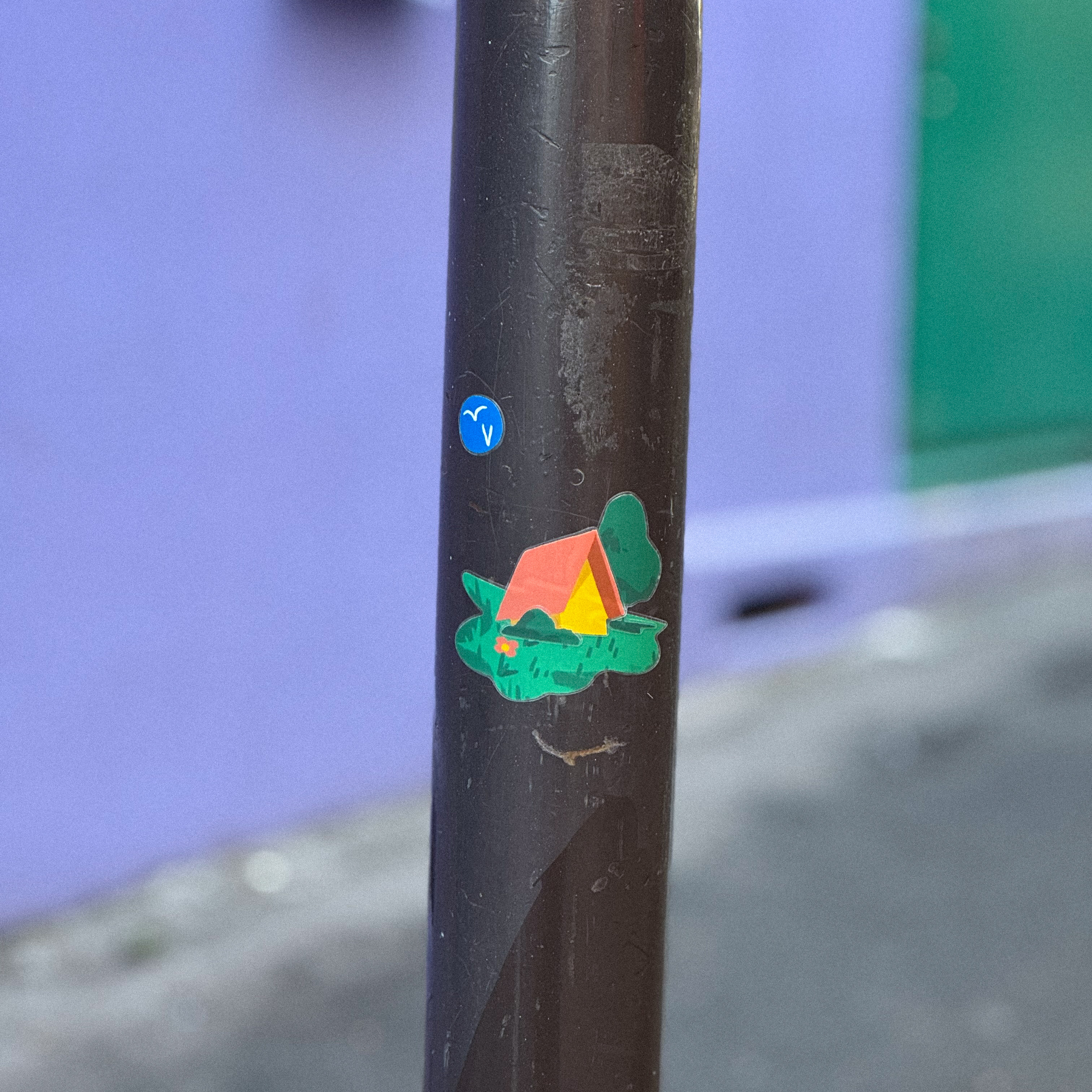 Transparent sticker of a small orange tent on green foliage, placed on a dark pole against a purple wall background.