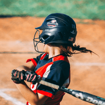 Softball player swings a bat on a dirt field, wearing a helmet with two vinyl stickers.