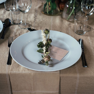 A table setting with a white plate, floral arrangement, name card, silverware, and glasses on a beige tablecloth.