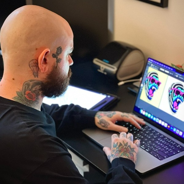 Tattooed artist working on a digital illustration of a roaring lion wearing sunglasses on a laptop screen.
